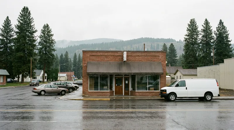 Independent community pharmacy storefront on a small Washington town street after rain, with evergreen trees and hills behind
