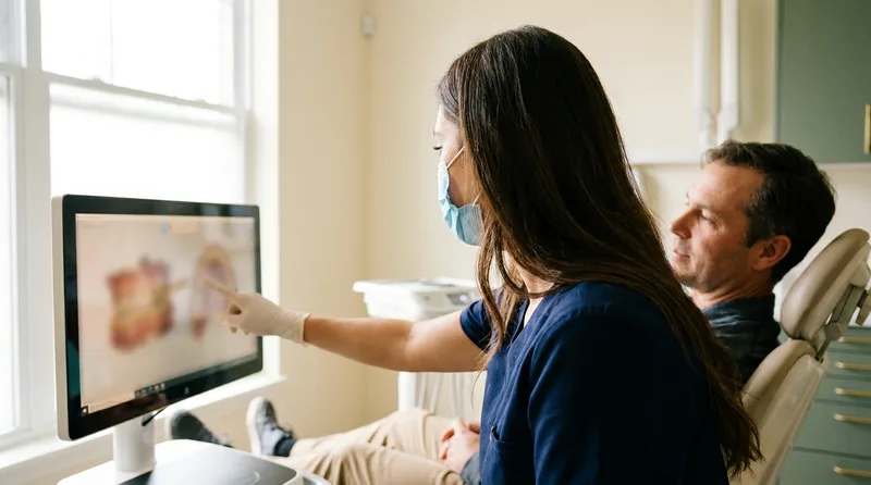 A dentist pointing to a 3D scan of a tooth and its roots on a monitor while a patient watches